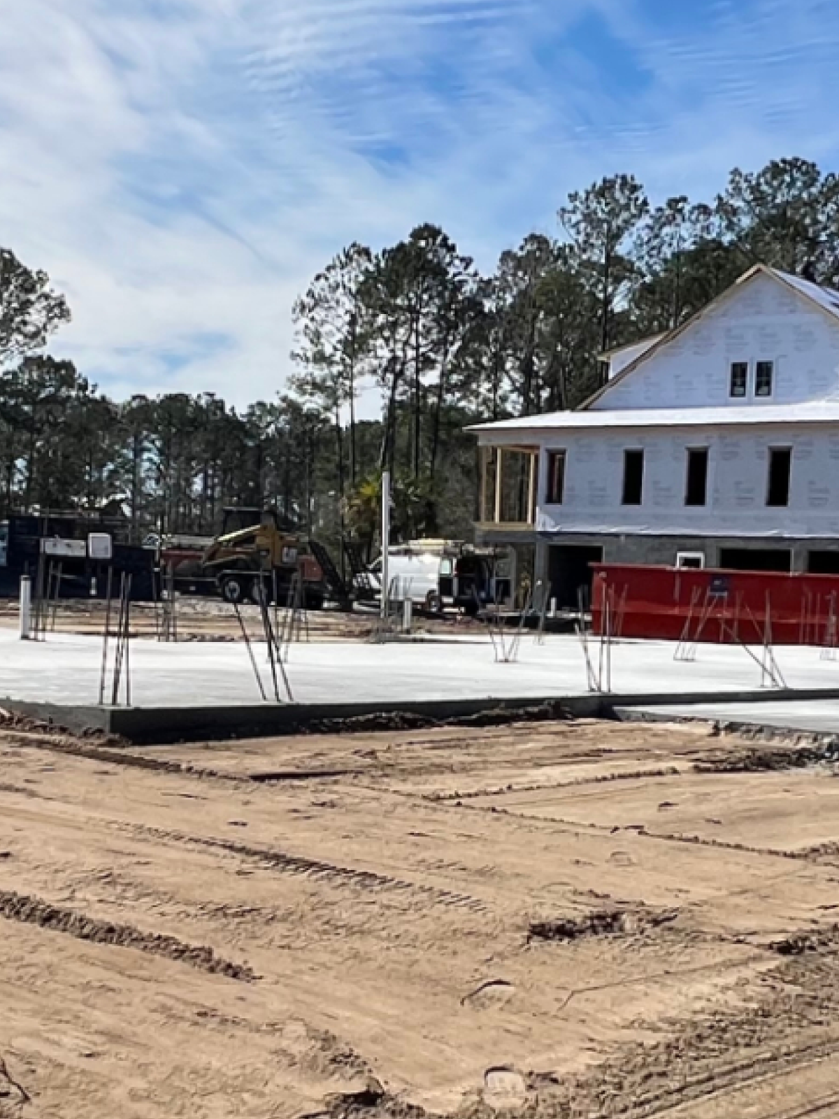 A house under construction with a concrete slab and exposed dirt, surrounded by pine trees under a blue sky.